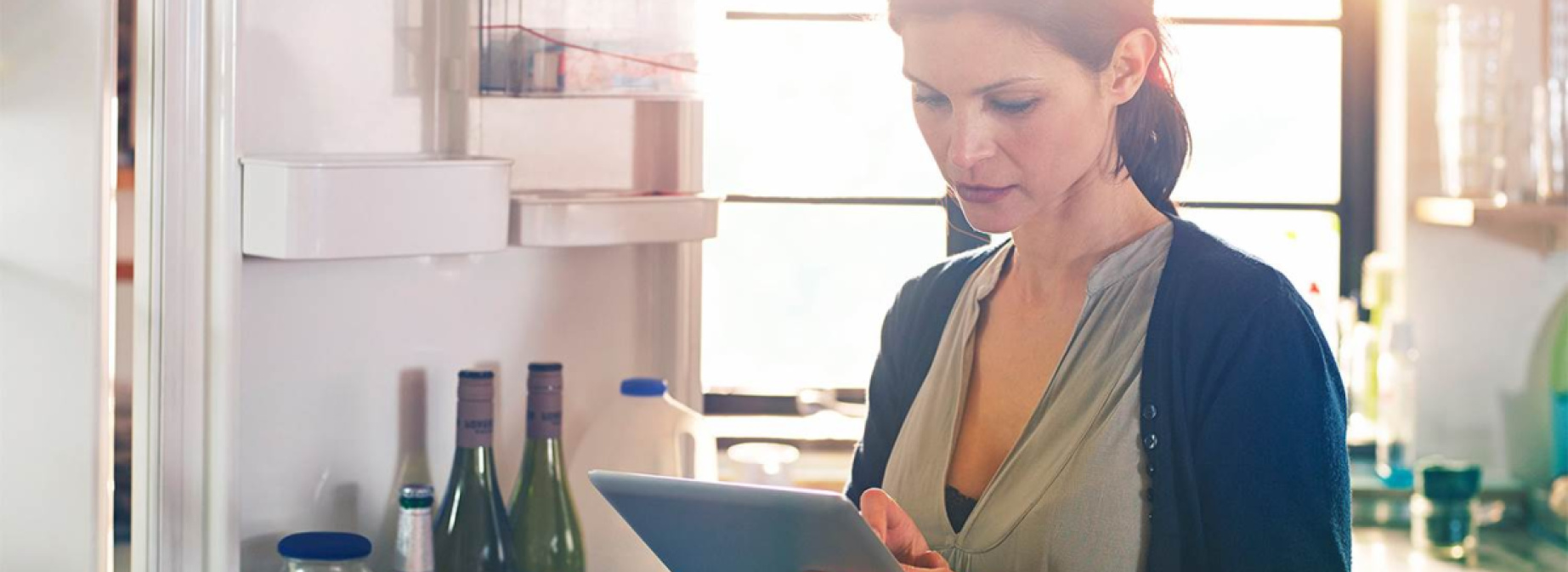 Woman is standing in front of open refrigerator and typing on her tablet