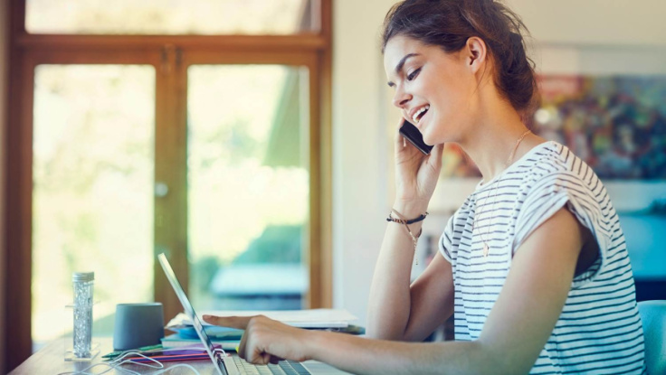 Young woman sitting at the desk and typing on her tablet while on the phone